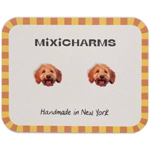 May include: A pair of stud earrings featuring a smiling, light brown dog face. The earrings are displayed on a cream-colored card with the text "MixiCharms" and "Handmade in New York" in black lettering, framed by a yellow and brown border.