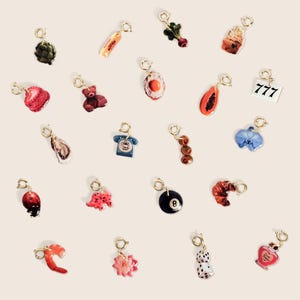 May include: A collection of gold-toned charms with various designs, including food items like a cupcake and shrimp, a teddy bear, a telephone, a lucky 777 sign, and a pair of dice. Each charm has a small clasp for attachment.