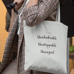 May include: A cream-colored tote bag with the words "Unshakable, Unstoppable, Uncaged" in black script. The bag is carried by a person wearing a plaid coat and holding a black umbrella.
