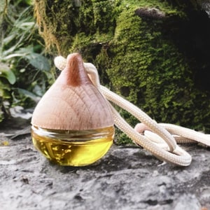 May include: A wooden and glass car air freshener with a light brown wooden top and a clear glass bottom filled with yellow liquid. A white rope is attached to the top. The air freshener sits on a gray stone surface.