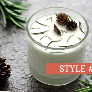 May include: A clear glass candle with white wax, decorated with sprigs of rosemary, small pinecones, and a wooden accent. The candle is labeled "STYLE #4" on a red banner. The candle sits on a textured gray surface.