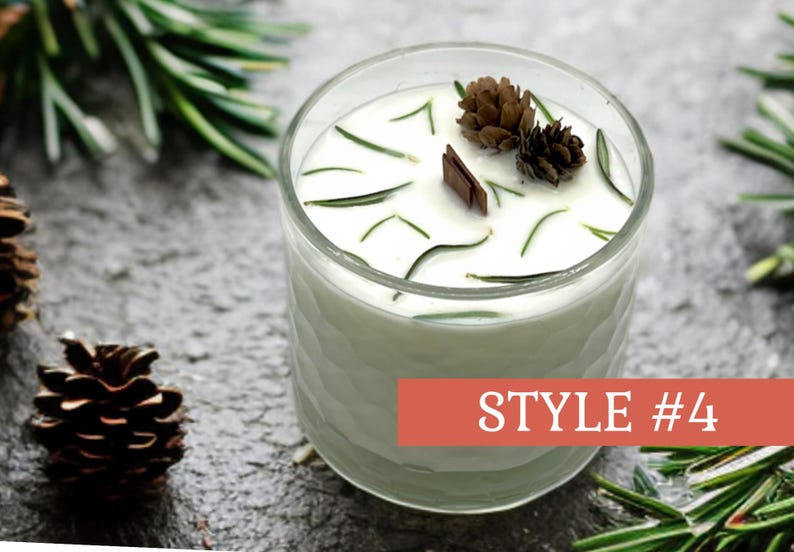 May include: A clear glass candle with white wax, decorated with sprigs of rosemary, small pinecones, and a wooden accent. The candle is labeled "STYLE #4" on a red banner. The candle sits on a textured gray surface.