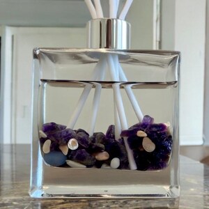 May include: A clear, square glass diffuser bottle with a silver cap and white reeds. The bottle contains a clear liquid and is filled with purple and white stones. The diffuser is on a marble surface.