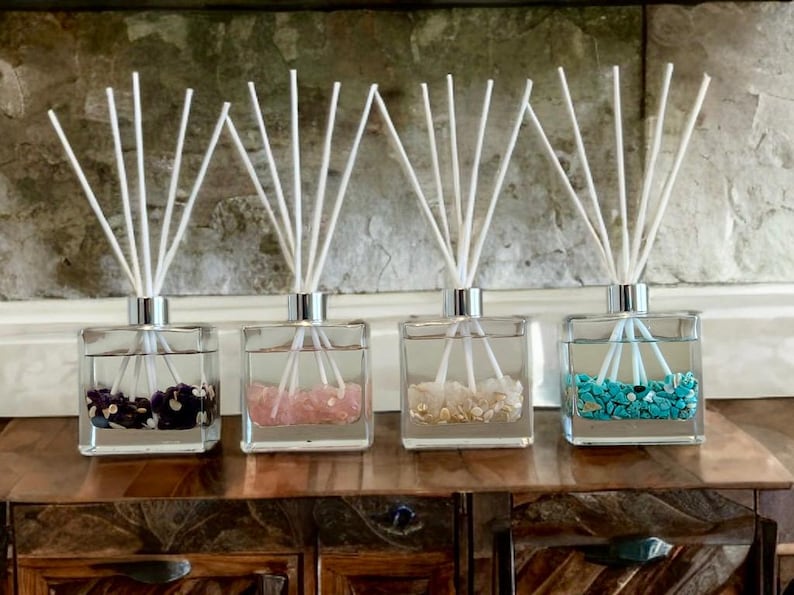 May include: Four square glass diffuser bottles with silver tops and white reeds. Each bottle contains colorful stones: purple, pink, beige, and turquoise. The bottles sit on a wooden surface.