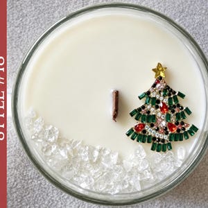 May include: A round candle in a glass container with smooth white wax and a single wooden wick. The surface features a decorative gold-tone metal Christmas tree brooch adorned with green, red, and clear crystals, plus clear crystal chips.