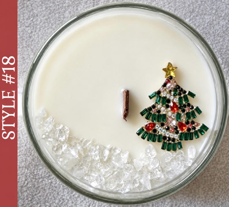May include: A round candle in a glass container with smooth white wax and a single wooden wick. The surface features a decorative gold-tone metal Christmas tree brooch adorned with green, red, and clear crystals, plus clear crystal chips.