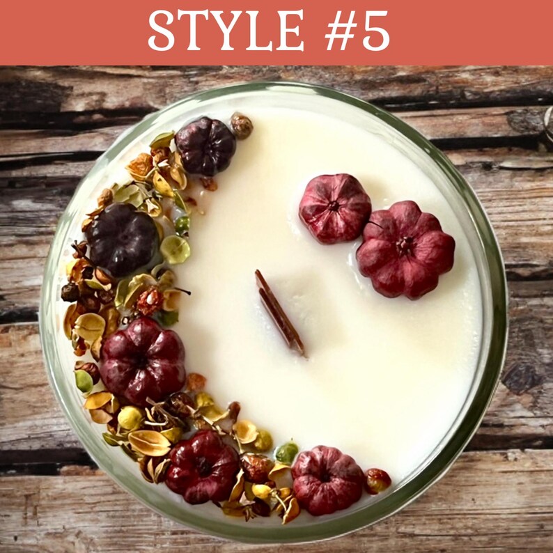 May include: A round candle in a clear glass container. The candle is white and decorated with burgundy-colored pumpkin-shaped accents, dried botanicals, and a cinnamon stick. The text "STYLE #5" is at the top.