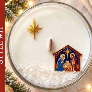 May include: A round glass candle container filled with white wax, featuring a central wooden wick, a gold-colored star charm, and a colorful enamel nativity scene charm. Clear crystal-like decorative stones are embedded in the wax surface.