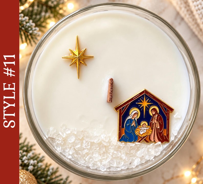 May include: A round glass candle container filled with white wax, featuring a central wooden wick, a gold-colored star charm, and a colorful enamel nativity scene charm. Clear crystal-like decorative stones are embedded in the wax surface.