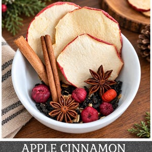 May include: A white bowl containing dried apple slices with red edges, two cinnamon sticks, two star anise, and small red berries mixed with dark tea leaves. The text APPLE CINNAMON is printed in white at the bottom of the image.
