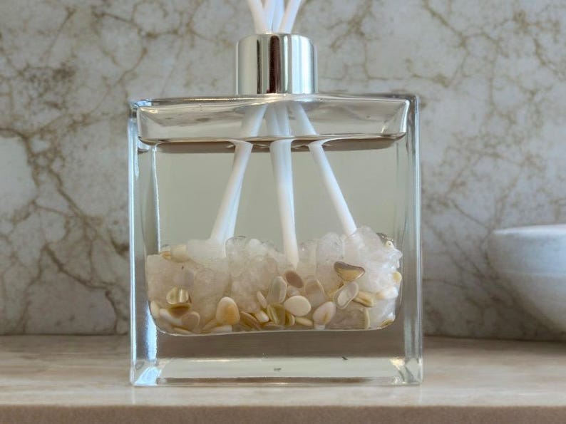 May include: A clear, square glass diffuser bottle with a silver cap and white reeds. The bottle contains clear liquid, white crystals, and small, tan, oval-shaped stones. The background is a marble-patterned wall.