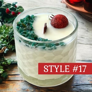 May include: A white candle in a clear glass container with a geometric textured surface. The candle features a wooden wick, a cluster of small green stones on one side, and a single red glittery spherical ornament. The text STYLE #17 is visible.