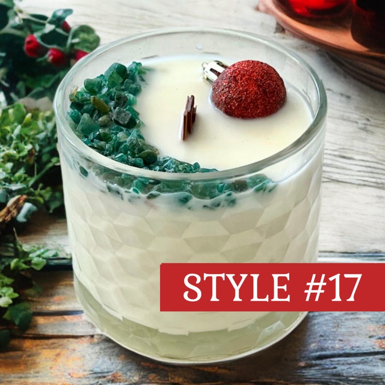 May include: A white candle in a clear glass container with a geometric textured surface. The candle features a wooden wick, a cluster of small green stones on one side, and a single red glittery spherical ornament. The text STYLE #17 is visible.