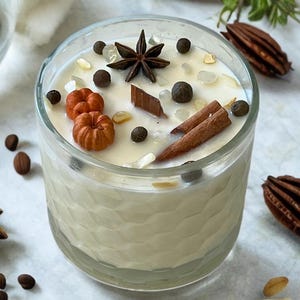 May include: A clear glass candle filled with creamy wax, decorated with star anise, cinnamon sticks, and small orange pumpkins. The candle is surrounded by scattered spices and nuts, creating a warm, inviting aesthetic.