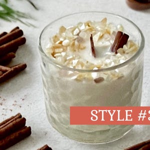 May include: A clear glass candle holder filled with a creamy wax and topped with decorative pieces and a wooden wick. Cinnamon sticks are arranged around the candle. The text "STYLE #3" is on a red banner.
