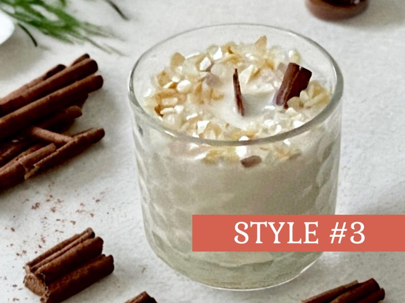 May include: A clear glass candle holder filled with a creamy wax and topped with decorative pieces and a wooden wick. Cinnamon sticks are arranged around the candle. The text "STYLE #3" is on a red banner.