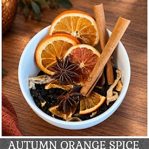May include: A white bowl filled with loose black tea leaves, dried orange slices, two brown cinnamon sticks, and two dark brown star anise pods. The text AUTUMN ORANGE SPICE is printed in white capital letters at the bottom of the image.