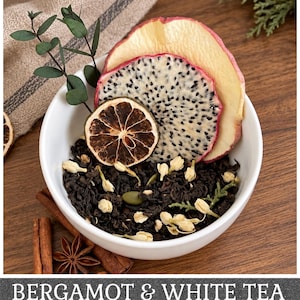 May include: A white bowl filled with loose leaf black tea, dried jasmine buds, a dried lime slice, a dried dragon fruit slice, and a dried apple slice. A sprig of eucalyptus is tucked into the tea. The text BERGAMOT & WHITE TEA is at the bottom.
