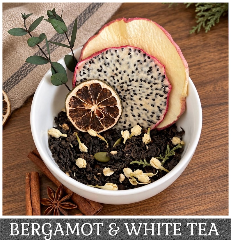 May include: A white bowl filled with loose leaf black tea, dried jasmine buds, a dried lime slice, a dried dragon fruit slice, and a dried apple slice. A sprig of eucalyptus is tucked into the tea. The text BERGAMOT & WHITE TEA is at the bottom.
