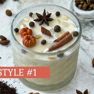 May include: A clear glass candle with a creamy wax interior, decorated with miniature orange pumpkins, star anise, cinnamon sticks, and peppercorns. The candle is labeled "STYLE #1" on a red banner.