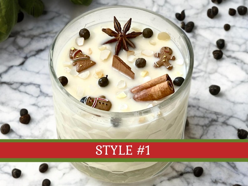 May include: A clear glass candle container filled with white wax, topped with decorative elements including a star anise, two cinnamon sticks, a chocolate piece, and small gingerbread-shaped figures, with the text STYLE #1 displayed on a red banner.