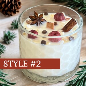 May include: A clear glass candle with a white wax interior, decorated with star anise, cinnamon, and small red and brown accents. The candle is labeled "STYLE #2" on a red banner. The candle sits on a wooden surface with pine needles and pine cones.