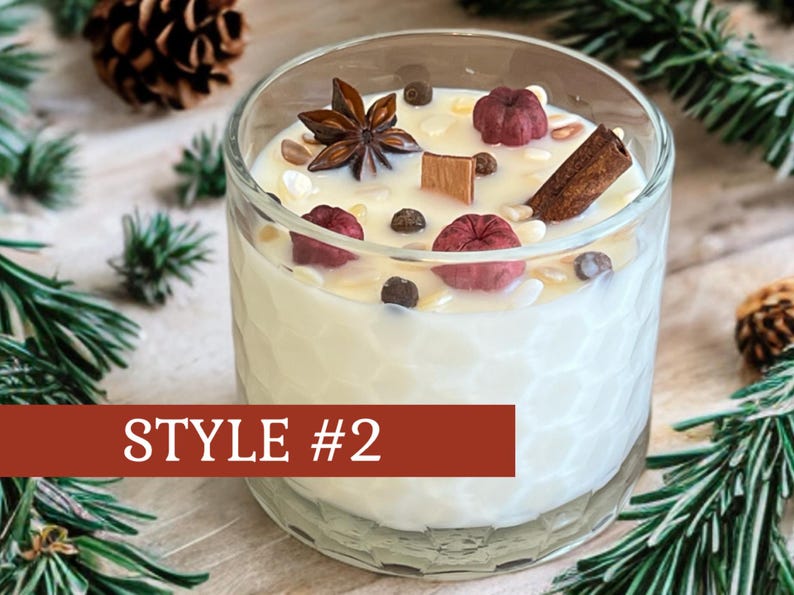 May include: A clear glass candle with a white wax interior, decorated with star anise, cinnamon, and small red and brown accents. The candle is labeled "STYLE #2" on a red banner. The candle sits on a wooden surface with pine needles and pine cones.