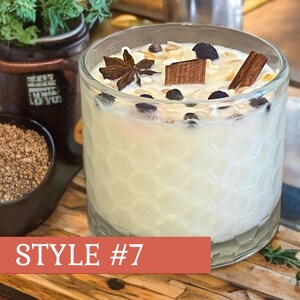May include: A clear glass candle with a white wax interior, decorated with star anise, cinnamon, and other toppings. The candle is labeled "Style #7" on a red banner. A small black bowl of brown granules is in the background.