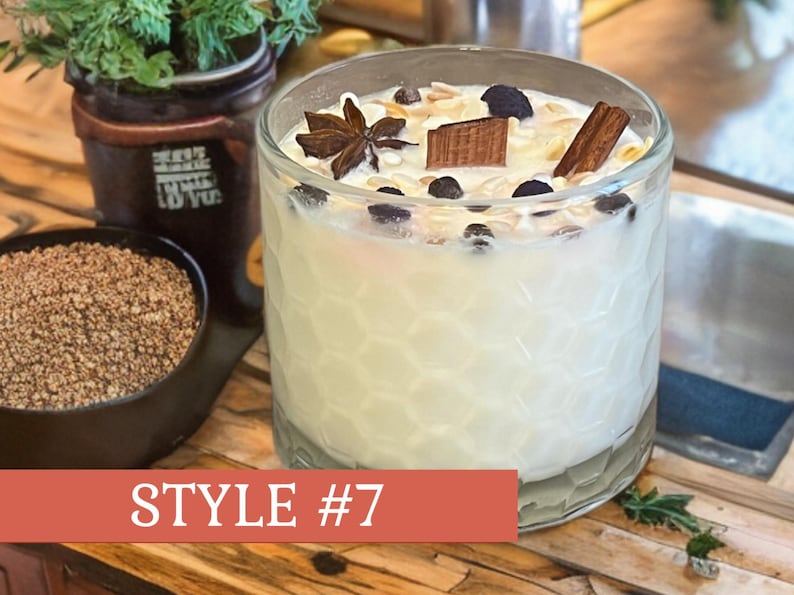 May include: A clear glass candle with a white wax interior, decorated with star anise, cinnamon, and other toppings. The candle is labeled "Style #7" on a red banner. A small black bowl of brown granules is in the background.