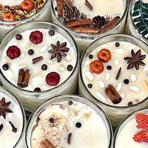 May include: A collection of glass jar candles featuring white wax topped with various decorative elements including star anise, cinnamon sticks, small wax pumpkins, dried leaves, orange slices, pinecones, and scattered dark brown beads.