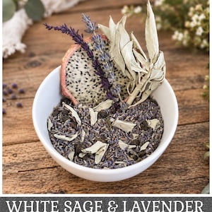 May include: A white ceramic bowl filled with a mixture of dried lavender buds, green tea leaves, and white sage pieces. A slice of dried dragon fruit, a sprig of purple lavender, and several white sage leaves are arranged upright in the bowl.