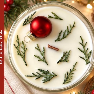 May include: A round candle in a glass container with white wax, featuring a single red ornament, several small green evergreen sprigs, and a small wooden wick centered on the surface. The text STYLE #2 is visible on the left side.