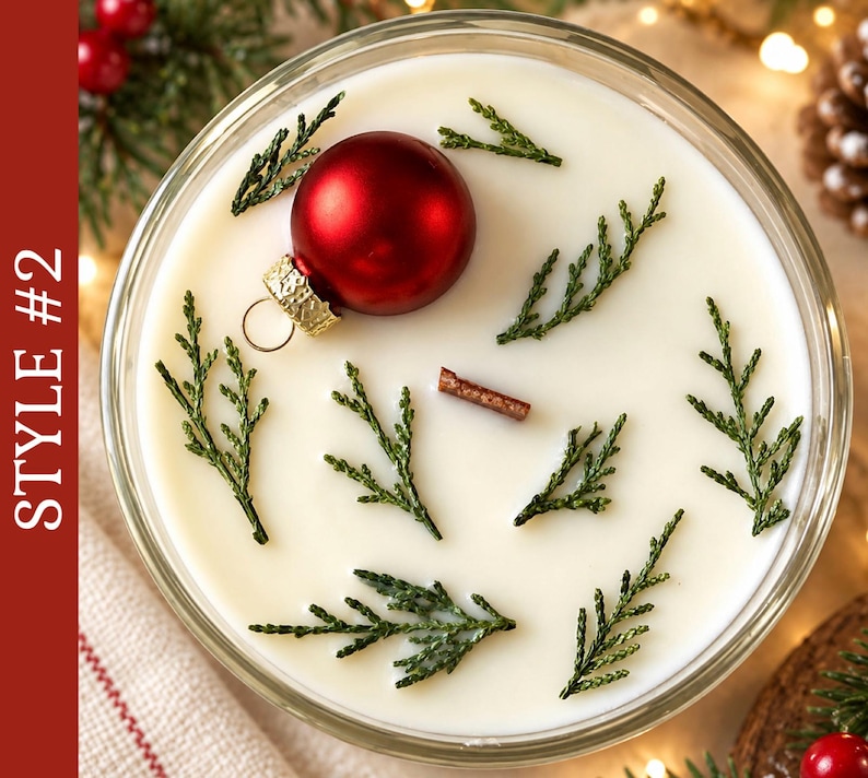 May include: A round candle in a glass container with white wax, featuring a single red ornament, several small green evergreen sprigs, and a small wooden wick centered on the surface. The text STYLE #2 is visible on the left side.