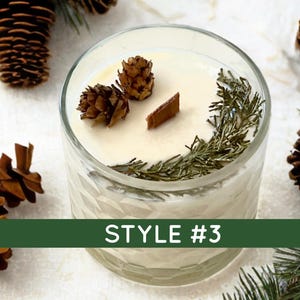 May include: A white candle in a clear, textured glass jar with a wooden wick. The wax surface is decorated with two small brown pinecones and a sprig of green evergreen needles. A green banner across the bottom displays the text STYLE #3.