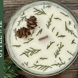 May include: A round glass candle container filled with white wax, featuring two small brown pinecones and scattered green evergreen sprigs on the surface. A single wooden wick is centered in the wax. The text STYLE #12 is visible on the left.