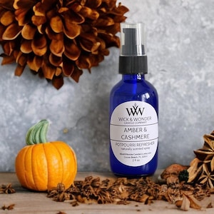 May include: A cobalt blue spray bottle with a black pump, labeled "Amber & Cashmere Potpourri Refresher." An orange pumpkin and dried spices are arranged around the bottle. The background features a large, dried flower.