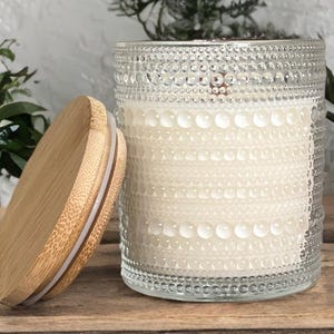 May include: A clear glass candle jar with a textured hobnail pattern and a cylindrical shape. The jar is filled with white wax and features a round, light-colored bamboo lid with a white silicone seal resting beside the glass container.