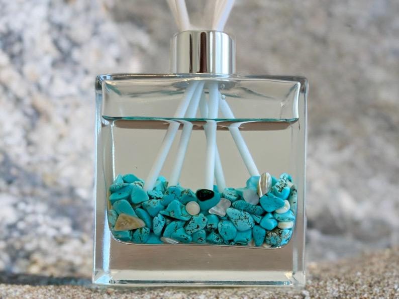 May include: A clear, square glass diffuser bottle filled with turquoise stones and white reeds. The bottle has a silver cap and is filled with a clear liquid. The reeds are arranged to disperse the fragrance.