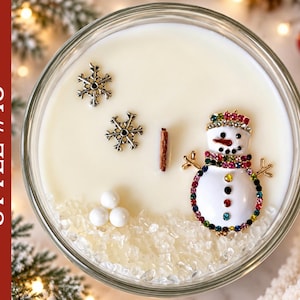 May include: A round glass candle container filled with white wax, featuring a wooden wick, two silver-toned snowflake charms, three small white spheres, and a snowman-shaped brooch decorated with colorful rhinestones. Text reads STYLE #13.