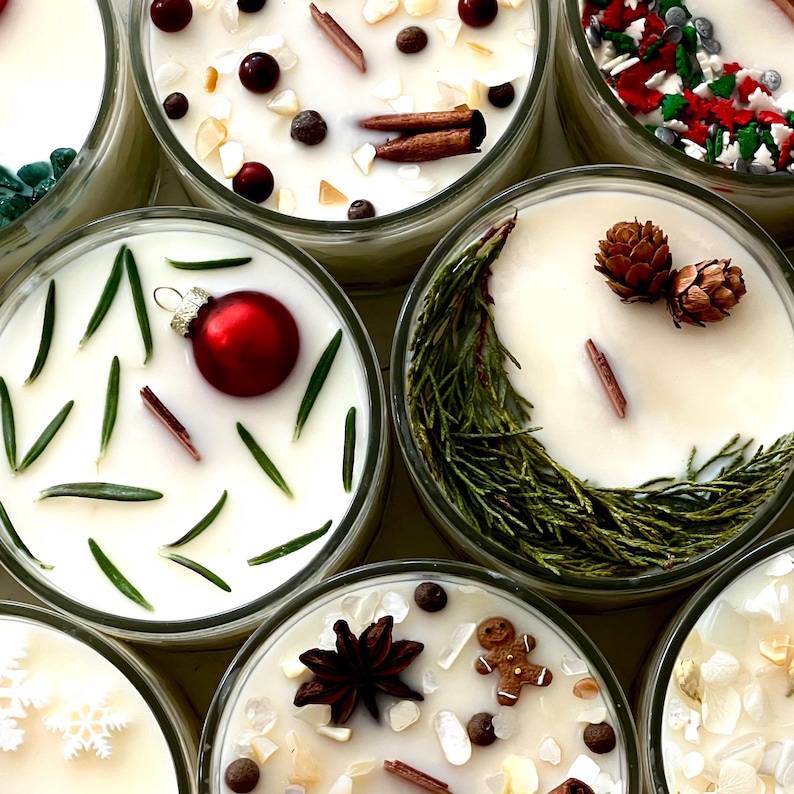 May include: Overhead shot of several glass jar candles with white wax and various decorative elements. Some candles have red ornaments, cinnamon sticks, and cranberries. Others feature pine cones, rosemary, and a gingerbread man, suggesting a holiday theme.