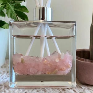 May include: A clear, square glass diffuser bottle with a silver cap, filled with a light pink liquid and rose quartz crystals. White reeds extend from the top. The diffuser sits on a white lace surface, with plants in the background.