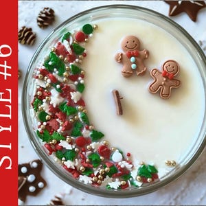 May include: A round glass candle container filled with white wax and a wooden wick. The surface is decorated with a crescent-shaped arrangement of red, green, and white holiday-themed sprinkles and gold beads, alongside two small gingerbread man figures.