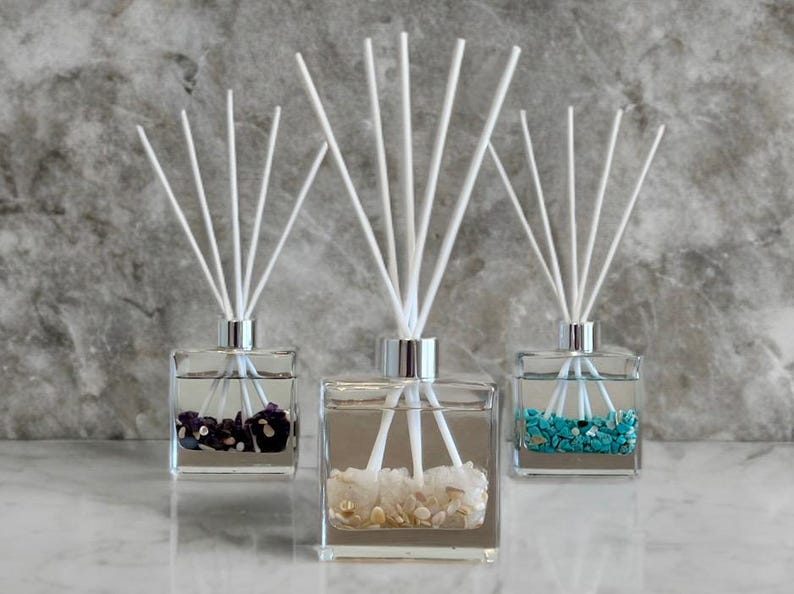 May include: Three glass reed diffusers with white reeds and silver collars. Each square bottle contains decorative stones in purple, white, and turquoise. The background is a mottled gray.