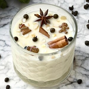 May include: A clear glass candle filled with creamy wax, decorated with gingerbread men, star anise, cinnamon sticks, and peppercorns. The candle sits on a marble surface, creating a festive and aromatic ambiance.