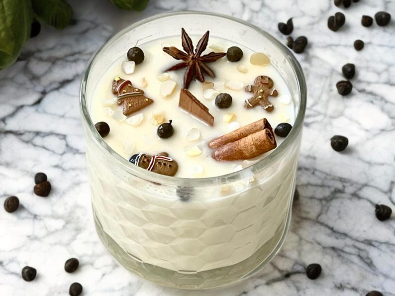 May include: A clear glass candle filled with creamy wax, decorated with gingerbread men, star anise, cinnamon sticks, and peppercorns. The candle sits on a marble surface, creating a festive and aromatic ambiance.