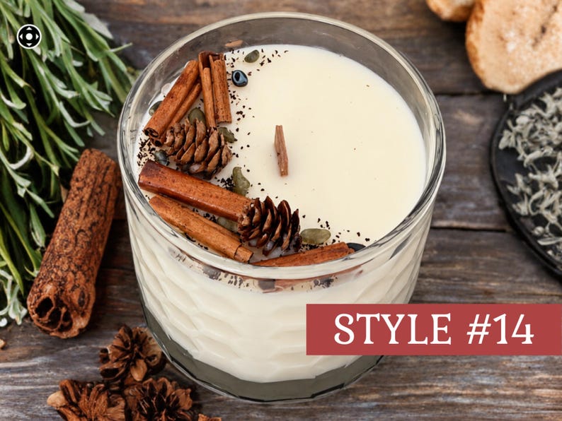 May include: A cream-colored candle in a clear, textured glass container with a wooden wick. The top is decorated with brown cinnamon sticks, small pinecones, and dark green botanical elements. A red rectangular label displays the text STYLE #14.