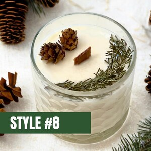 May include: A clear glass candle with a cream-colored wax, decorated with small pine cones, sprigs of greenery, and a wooden wick. The candle is labeled "STYLE #8" on a green rectangular label.