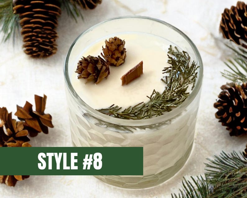 May include: A clear glass candle with a cream-colored wax, decorated with small pine cones, sprigs of greenery, and a wooden wick. The candle is labeled "STYLE #8" on a green rectangular label.