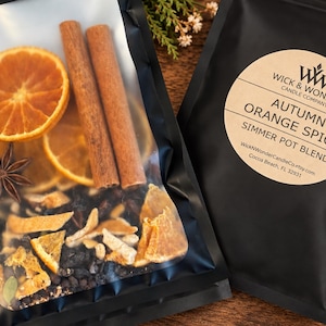 May include: A black pouch containing a simmer pot blend of dried orange slices, two cinnamon sticks, and a star anise. The label reads WICK & WONDER CANDLE COMPANY, AUTUMN ORANGE SPICE, SIMMER POT BLEND, and includes the website and location address.