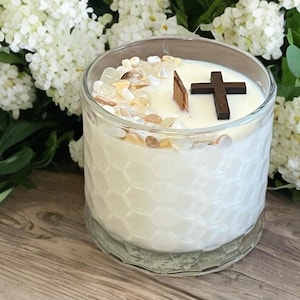 May include: A white candle in a clear glass jar with a honeycomb pattern. The candle is topped with small stones and a dark wooden cross. The background features white flowers and green leaves.